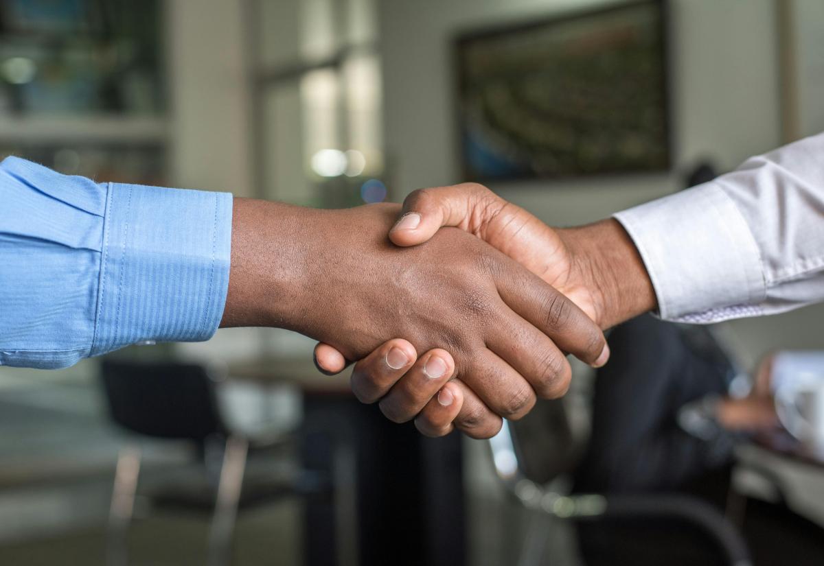 Two hands shake, both wearing shirts. An office is blurred in the background.