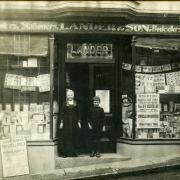 A mounted photograph showing shop front with posters announcing events in Ruan Minor, Mullion, Helston and Porthleven. Lady and boy stood on doorstep. Lander & Son, Printers, Stationers, Booksellers and Newsagents