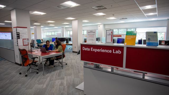 Photo shows a red sign with white lettering indicating the entrance to the "Data Experience Lab," a computing lab with an open and airy design. At the forefront of the image to the left of the sign is a table at which a data science consultant sits with a computer.  She is providing assistance to another student. Behind them, a large wall of windows lights the features of the space. Around the room are computer workstations, a seating area with whiteboard tables, and various data visualizations.