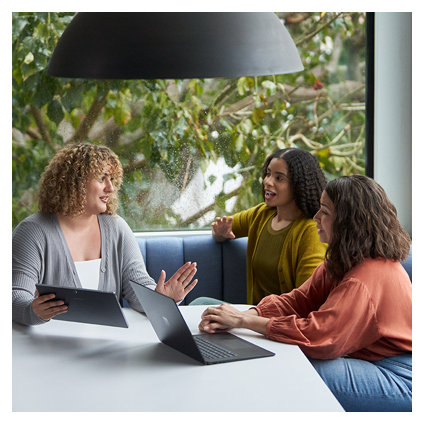 Three female coworkers sitting at a table talking and while looking at a Surface computer and tablet