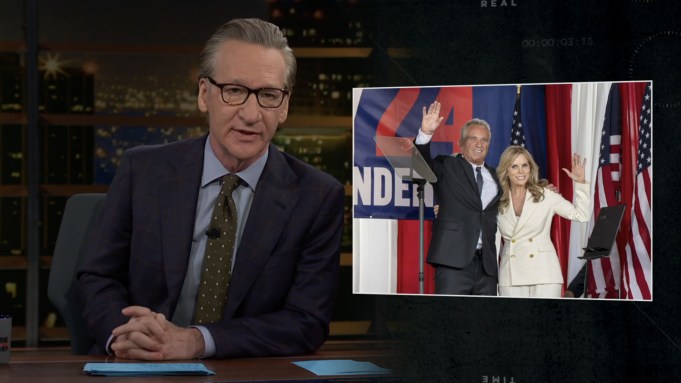 A grab of Bill Maher sitting at his 'Real Time' desk and speaking with an image displayed next him of Robert F. Kennedy Jr. and wife Cheryl Hynes.