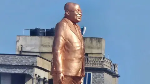 The statue of Nana Akufo-Addo, which is bronze in colour. He is depicted wearing a suit, his glasses and is smiling. It is outside Effia-Nkwanta Regional Hospital in Sekondi, Ghana - November 2024