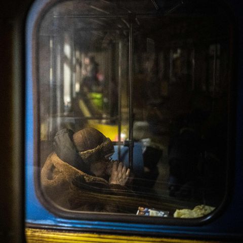 Hinter einem schmutzigen Fenster einer blau-gelb lackierten U-Bahn sitzt eine alte Frau im Wintermantel und trinkt Tee