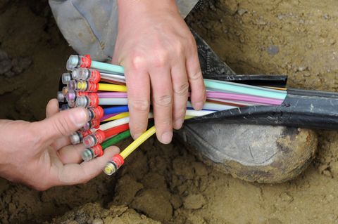 Ein Bauarbeiter kniet in einem Erdloch und hält in seinen Händen einen Strang mit unterschiedlich farbig markierten Kabeln