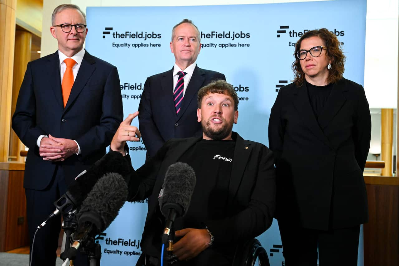 A  man in a wheelchair speaks into a microphone. Two men and a woman are behind him.