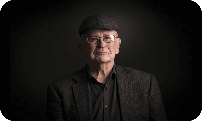 Photographic portrait of an older man in a dark tweed jacket and cheesecutter hat. Beautifully lit so that only his face is really visible. An air of great warmth and cleverness.