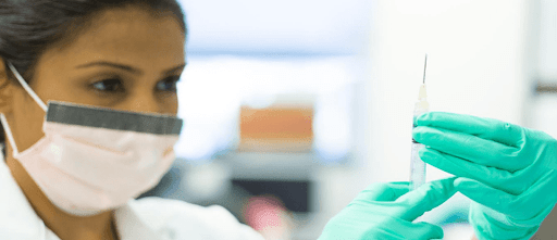 Woman with surgical mask, lab coat, and gloves holds up a syringe at eye level