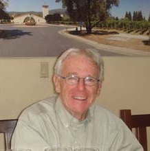 A man with white hair and glasses wearing a brown button-down shirt, smiling at the camera