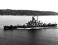 A battleship sails on calm waters with land in the background.