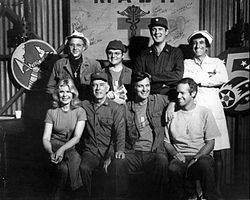 El repartiment de M*A*S*H de la temporada 6, 1977 (en el sentit horari de l'esquerra): William Christopher, Gary Burghoff, David Ogden Stiers, Jamie Farr, Mike Farrell, Alan Alda, Harry Morgan i Loretta Swit.