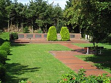 Frontale Farbfotografie eines Gedenkorts auf einem Friedhof. Mehrere graue Tafeln mit viel Text sind an einer flachen Mauer angebracht. Vor der Mauer sind zwei schmale Hecken mit einem Gedenkstein in der Mitte. Von dem Gedenkort führt ein braun gepflasterter Weg an einer Bank mit Baum vorbei und endet in der rechten unteren Bildecke. Der Rest ist mit Rasen ausgestattet und im Hintergrund stehen Bäume.