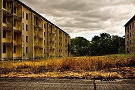 Une barre d'immeuble décrépie à chaque côté de l'image. Devant l'objectif, une route debéton et de l'herbe haute.