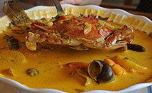 Photo of a bowl of tapado soup, yellow-orange in color, with crab, fish, and other ingredients visible in the bowl