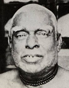 A close-up portrait of an old man with grey hair and thick wooden necklace beads