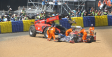 A race car being removed from the circuit after an accident by five people wearing orange for visibility