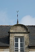 Photographie d'une lucarne surmontée d’une girouette, sur fond de toit en ardoise.