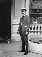 Black and white image of a man in a suit and Cordovan hat, smoking a cigarette and standing in front of a column and a window in front of a building with a dog emerging on the left from the entry door.