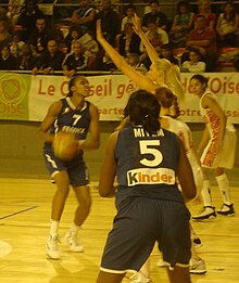 Au centre, joueuse en bleu se préparant à faire une passe à une joueuse au premier plan, de dos. Face à la première des joueuses en blanc lèvent les bras.