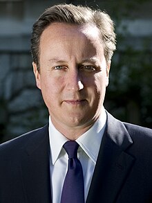 A man with dark hair and blue eyes, wearing a navy suit with a blue tie as well as smiling and facing to his right.