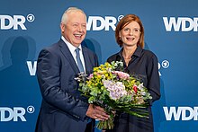 Tom Buhrow (links) überreicht Katrin Vernau (rechts) einen Blumenstrauß vor blauer Wand,