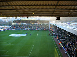 The inside of an association football stadium, with a stand on the right-hand side full of supporters. The pitch is visible to the left of the stand, with a floodlight in the background.