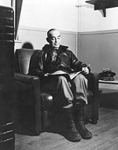 A man wearing a bomber jacket and ski boots sitting in an armchair, reading.