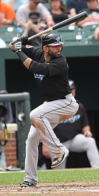 A man in a black batting helmet, black baseball jersey, and grey pants holds a baseball bat midway into right-handed swing with his left leg up in the air.