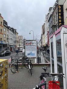 Straßenszene in Köln mit einem Plakat von Dies Irae über Korruption bei der FIFA