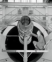 photo noir et blanc. moteur installé sur une plate forme, devant un tunnel de soufflerie. Un technicien, accroupi, l'inspecte.