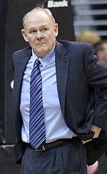 A man, wearing a black suit and a blue tie, is watching the basketball game while sitting courtside.