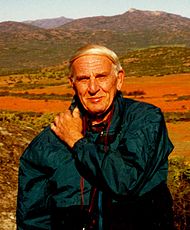 Edward M. Burgess pictured in Namaqualand, South Africa. Burgess is wearing a dark blue-green jacket and is carrying a camera strapped around his neck (tourist style). His left arm is positioned across his chest with his left hand on his right shoulder. Burgess is looking directly into the (active) camera with a slight smile. In the background are numerous mountainous peaks breaking to a pale blue sky at the top of the photo. At approximately shoulder height the green vegetation in the foot hills gives way to an expanse of orange-red flowers.