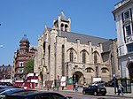 A street sloping uphill from right to left. There are parked cars and pedestrians in the foreground, and the hoardings further up indicate that the road is temporarily closed. On the right of the road in the centre of the picture is one end of a large early twentieth-century stone-built church in the Arts and Crafts style which is apparently being restored, as there are portakabins and screens outside. It has an elaborate entrance on the street with a large recessed window above and another to the right. There is another, plainer, entrance on the minor street with a No Entry sign that runs alongside the side of the church. Alongside it are two small windows; above and behind this entrance and the windows are three large arches with, underneath each, a tall thin window. Beyond these, part of a transept can be seen. Above the church is a bell-tower with castellated columns at the corners and a pyramid roof. There are railings between the church and pavement. Beyond the church, some red-brick buildings can be seen, most notably a four-storey building with an elaborate corner tower. In the right foreground, across the side-street from the church, part of a four-storey 1920s neo-Georgian building can be seen behind a pair of telephone kiosks.