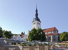 Frontale Farbfotografie von einem weißen Kirchturm mit dunkler Turmlaterne und einem Kirchenschiff im gotischen Stil. Das Kirchenschiff hat eine graue Steinfassade und ein rotes Dach. Eine Mauererhöhung mit Treppen führt zur vorderen Grünanlage.