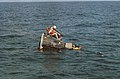 Photographie couleur d'une capsule Gemini dans l'eau avec les deux astronautes et un plongeur visibles.