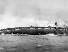 Una grande nave da guerra si sta rovesciando sul fianco, i marinai affollano il bordo emerso della nave, aggrappandosi disperatamente alle reti antisiluro. Sullo sfondo un'enorme nube di vapore si leva dal relitto.