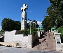 Farbfotografie von einem großen, weißen Steinkreuz auf der linken Seite einer Mauererhöhung. Rechts führt eine Treppe mit altern Laternen hinunter und an beiden Seiten sind Alleen gepflanzt.