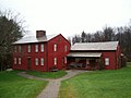 A red colonial house with a large central chimney stands at the end of a dirt path. A second building is connected near one corner, in front of which is a covered porch with benches.