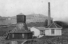 Photo en noir et blanc de petits bâtiments, avec grande cheminée et tour pyramidale en bois fermée au pied de la colline de la chapelle Notre-Dame-du-Haut.