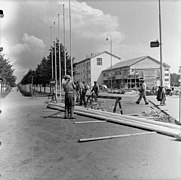 Une photo en noir et blanc représentant une place, une équipe de construction portant des poutres, et un bâtiment en fond, ainsi que des lampadaires déjà installés.