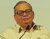 A photograph of a speaking man with spectacles in front of a mic