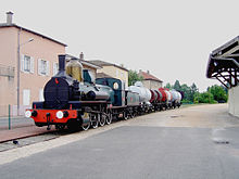 La photo couleur représente une locomotive à vapeur bleu attelée à des wagons citernes et bifoudres.