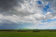 Photographie d'une plaine plate avec des champs au premier plan, des bois en second plan, et avec un ciel nuageux.