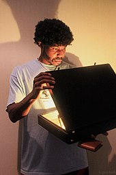 Un homme de race noire avec une coiffure afro vêtu comme le personnage de Jules Winnfield à la fin du film et ouvrant une mallette qui produit une forte lueur.