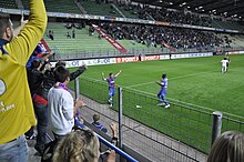 Joueur célébrant son but devant ses supporters, ceux-ci étant debout.