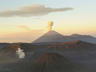 Un volcán marrón en el centro con humo blanco emanando de la cumbre, un cielo nublado que se convierte en azul en azul en cima de la cumbre y en amarillo en el medio al rojo en el horizonte, y montañas de color marrón en el primero plano.