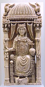 A carving of a crowned woman seated on a throne behind undrawn curtains. She is holding a cross atop a globe on her left hand