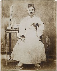 Photographie en noir et blanc montrant un homme tenant un livre et assis à côté d’une petite table.