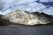 Daytime view of a large body of water standing before a prominent peak, which communicates with several others partly out of view and behind. A gravel beach at the far end of the lake gives way to steep slopes leading up to the peaks; The mountains lack trees. Patchy snowcover defines their recesses, and whitish vein-like streaks extend up from the base of the largest.