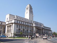 A monumental twentieth-century neo-classical building of Portland stone stands on a main road with blue sky and clouds behind and above it. It has a basement and four floors, with a flight of steps leading up to a recessed portico and above it a tall clock-tower in four stages in Greek revival style. A lower addition extends out of the picture to the left. In front is an island flower-bed and on the other side of the road are a number of older brick buildings. A number of pedestrians, a bus and a taxi are also to be seen, as well as a left-pointing sign saying "Alternative hgv route to the University".