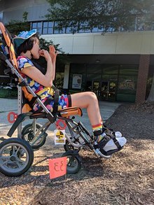 A girl with short brown hair sits in a neon orange push chair. She is on a concrete pathway in front of a building.